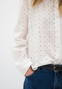 Camicia bianca con bottoni, motivo a griglia e fori intagliati. Maniche lunghe e polsini, indossata con jeans in denim blu e una cintura nera.