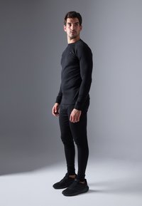 Top negru cu mânecă lungă și pantaloni strâmți, ambele dintr-un material neted. Modelul poartă pantofi sport negri, stând în fața unui fundal gri.