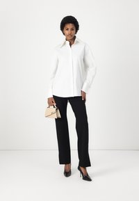 White button-up shirt with a classic collar, paired with black wide-leg trousers. Holds a small beige handbag with a signature. Black heeled shoes.