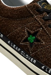 Brown fuzzy sneaker with black laces, featuring a green embroidered four-leaf clover inside a star. Black and white logo on the sole.