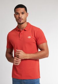 Červené bavlněné polo tričko s knoflíkovou légou na třech knoflících, krátkými rukávy a malou nášivkou loga na hrudi. Nosené s modrými džíny.