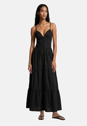 Donna che indossa un vestito lungo nero senza maniche con bottoni sulla parte anteriore, collana d'oro, bracciale e sandali neri.