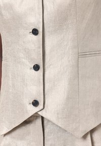 Beige linneväst med en figursydd design, sex svarta knappar och en spetsig nederkant. Inkluderar diskret struktur med sidofickor.