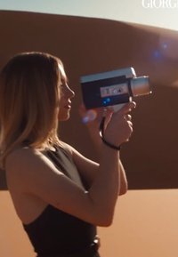 Femme en robe noire filmant avec une caméra vidéo vintage devant un paysage désertique pendant l'heure dorée.