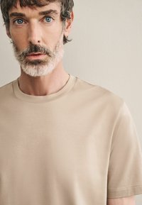 Púder színű, rövid ujjú póló sima anyagból, amely egyszerű környakú fazonnal és finom varrási részletekkel rendelkezik.
