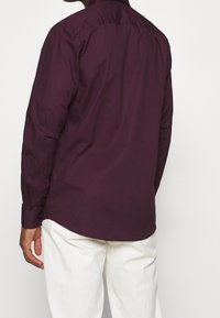 Lange mouwshirt in donkerpaarse stof met een ronde zoom en knopenmanchetten, gecombineerd met witte broek. Heeft een rugpas en kraag.