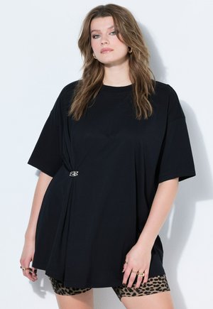 Ung kvinde med bølget hår bærer oversize sort t-shirt spændt sammen med klemme og leopardmønstrede cykelshorts mod hvid baggrund.