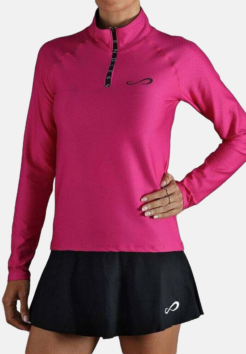 Vrouw in een felroze langemouwen, half rits bovenstuk en een korte zwarte rok met een wit logo op de zoom.