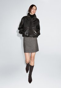 Schwarze Leder-Bomberjacke mit zwei Fronttaschen, grauer Woll-Minirock und braune knöchellange Socken, kombiniert mit spitz zulaufenden schwarzen Schuhen.