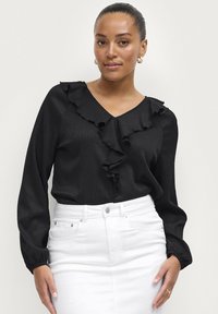 Blusa nera con scollatura e polsini arricciati, realizzata in tessuto testurizzato. Indossata con una gonna bianca a vita alta, che mette in evidenza una silhouette aderente.