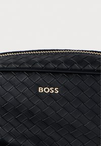 Czarna pleciona skórzana kopertówka z złotymi elementami metalowymi, wyposażona w zamek błyskawiczny i tłoczone logo marki z przodu dla dodatkowego efektu.