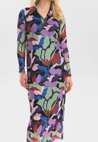 Kvinna klädd i en långärmad, kragad maxiklänning med ett flerfärgat abstrakt blommönster i lila, grönt, blått, svart och rött.