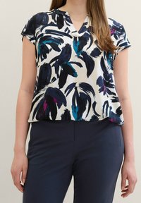 Blusa a maniche corte con scollo a V. Presenta una base bianca con modelli floreali astratti in blu navy, teal e viola. Tessuto leggero, vestibilità comoda.
