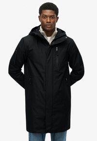 Zwarte waterdichte jas met een capuchon, voorzak met rits en zijritsen, met een gladde textuur en een lang ontwerp. Draagt over een lichte trui.