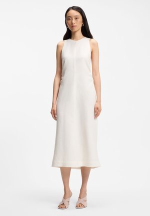 Femeie în picioare, purtând o rochie midi fără mâneci, albă, și pantofi cu toc deschis, roz deschis, pe un fundal alb simplu.