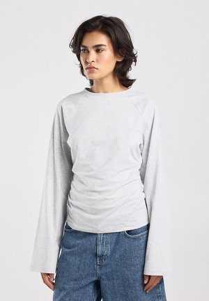 Jonge vrouw met kort donker haar, gekleed in een lichtgrijs shirt met lange mouwen en blauwe spijkerbroek, kijkt iets naar de zijkant tegen een eenvoudige achtergrond.