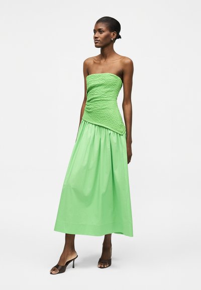 Γυναίκα που φοράει πράσινο τοπ χωρίς τιράντες με υφή και ταιριαστή ψηλόμεση, μακριά φούστα με μαύρα σανδάλια με τακούνι, στέκεται μπροστά σε απλό φόντο.