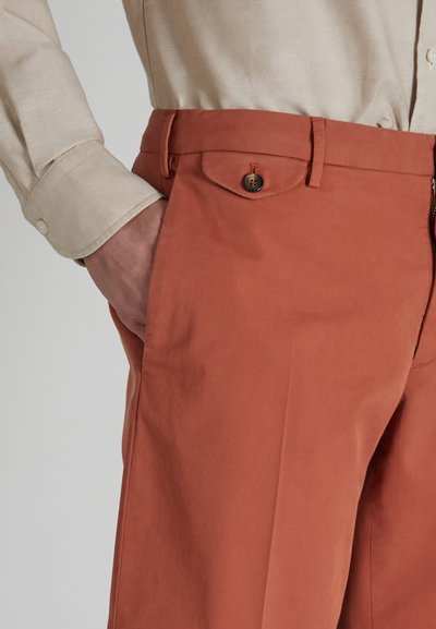 Pantaloni color ruggine realizzati in tessuto liscio, con chiusura a bottone e tasche laterali. La vita ha un design sartoriale.