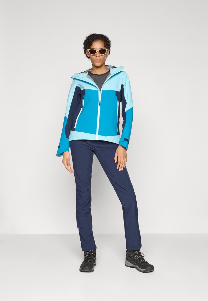 Helles blau und dunkelblaues Zip-Jackett mit Kapuze, ausgestattet mit elastischen Bündchen. Getragen mit dunklen Hosen und schwarzen Wanderschuhen, ergänzt mit Sonnenbrille.