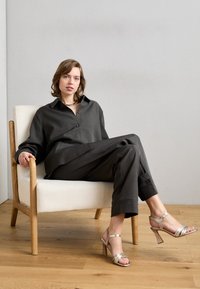 Camisa de botón gris de gran tamaño y pantalones a juego, combinados con tacones plateados de tiras, sentada en una silla de marco de madera con un cojín blanco.