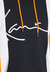 Marinblå hoodie med orange dragsko och vit broderad logotyp. Har kontrasterande vita och orange sidopaneler. Mjuk tygkänsla.