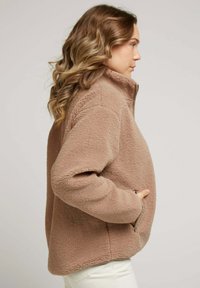 Pull en polaire marron avec un col montant, fermeture éclair demi-zip sur le devant et poches latérales. Présente un tissu texturé et une coupe décontractée, avec des manches longues.