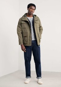 Parka vert olive avec capuche, plusieurs poches à l'avant, porté par-dessus une chemise rayée, associé à un jean sombre et des baskets blanches.