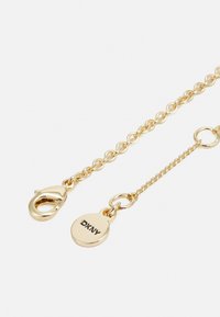 Guld kedja armband med en kräftklipp och en tagg som har "DKNY." Kedjan har en vriden textur med en slät yta.