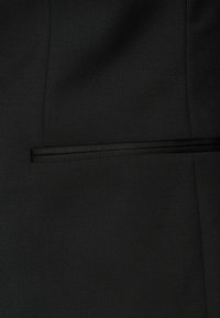 Svart tyg med en slät, texturerad yta. Inkluderar en tunn, horisontell svart tejpdekoration över fickområdet.