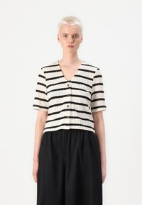 Pletený cropped top s vertikálními černými pruhy na bílém pozadí, výstřihem do V, krátkými rukávy a dřevěnými knoflíky uprostřed přední části.