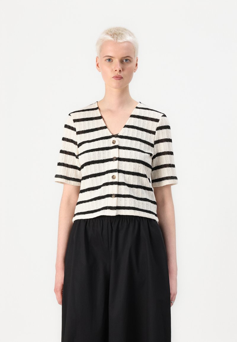 Pletený cropped top s vertikálními černými pruhy na bílém pozadí, výstřihem do V, krátkými rukávy a dřevěnými knoflíky uprostřed přední části.