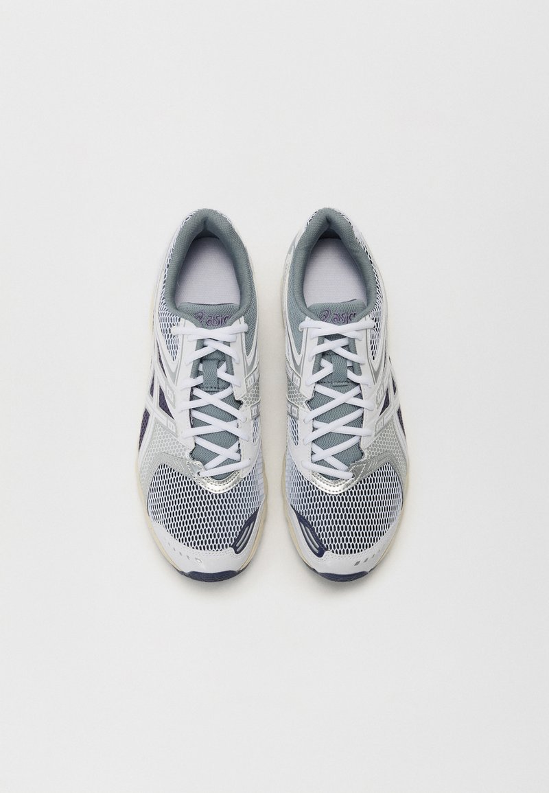 Zapatillas deportivas blancas y grises con parte superior de malla, cierre de cordones y acentos morados. Presentan una suela texturizada para agarre y soporte.