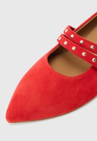 Piros velúr lapos cipő hegyes orral, két díszítő pánttal, melyeket fém szegecsek díszítenek. Simább textúra és letisztult kialakítás.
