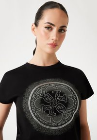 Black T-shirt featuring a circular design with intricate metallic silver accents and rhinestones in a patterned arrangement. Smooth texture.
