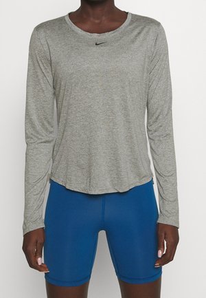 Persoon die een grijze lange mouw Nike-top en blauwe strakke sportshort draagt, staand tegen een effen witte achtergrond.