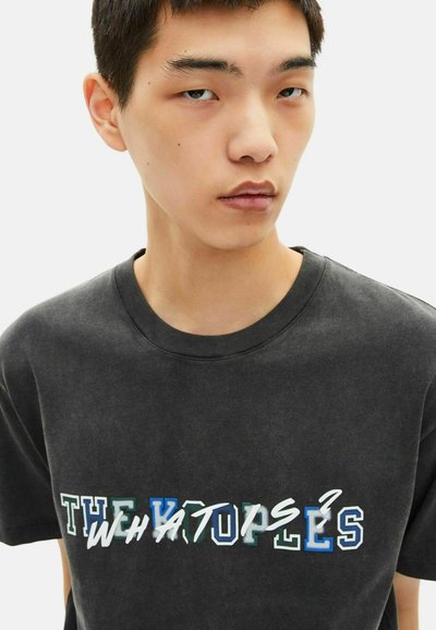 Juoda medvilninė marškinėliai su apvaliu kaklu, puikuojanti ryškiu, daugiaspalviu tekstu „THE WHATOPS?“ ant priekio. Lygus paviršius.