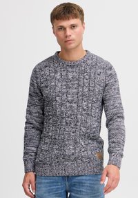 Young man wearing a grey and black patterned cable knit sweater and blue jeans against a plain light background.