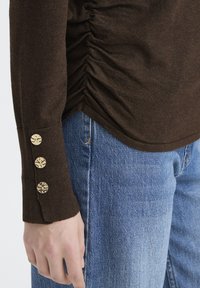 Pruun pika varrukaga särk külgse ruššimise ja kolm kuldset dekoratiivset nööpi mansetil, kombineeritud helesiniste teksapükstega.