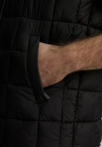 Czarna kamizelka puchowa z teksturowanym, pikowanym wzorem i boczną kieszenią. Materiał wydaje się lekki i izolujący.