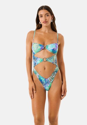 Donna che indossa un costume intero multicolore con stampa animalier e ritagli, con dettagli ad anello sul davanti, in piedi davanti a uno sfondo semplice.