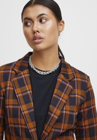 Rutet blazer i orange, marineblå og lilla nuancer, med skarpe revers og en skræddersyet pasform, lagd over en sort t-shirt med en sølv kæde halskæde.