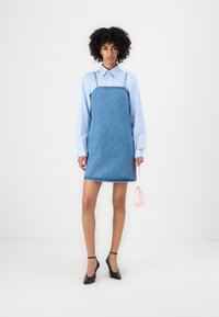 Denim overall dress dengan tali yang dapat disesuaikan, kain biru, hem kasar, dipakai di atas kemeja berkerah biru muda, dipadukan dengan sepatu hak hitam dan tas kecil berwarna pink.