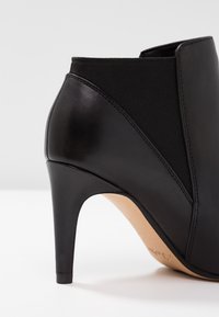 Botín negro con un exterior de cuero elegante y panel elástico. Presenta un alto tacón de aguja y un acabado texturizado a lo largo de los costados.