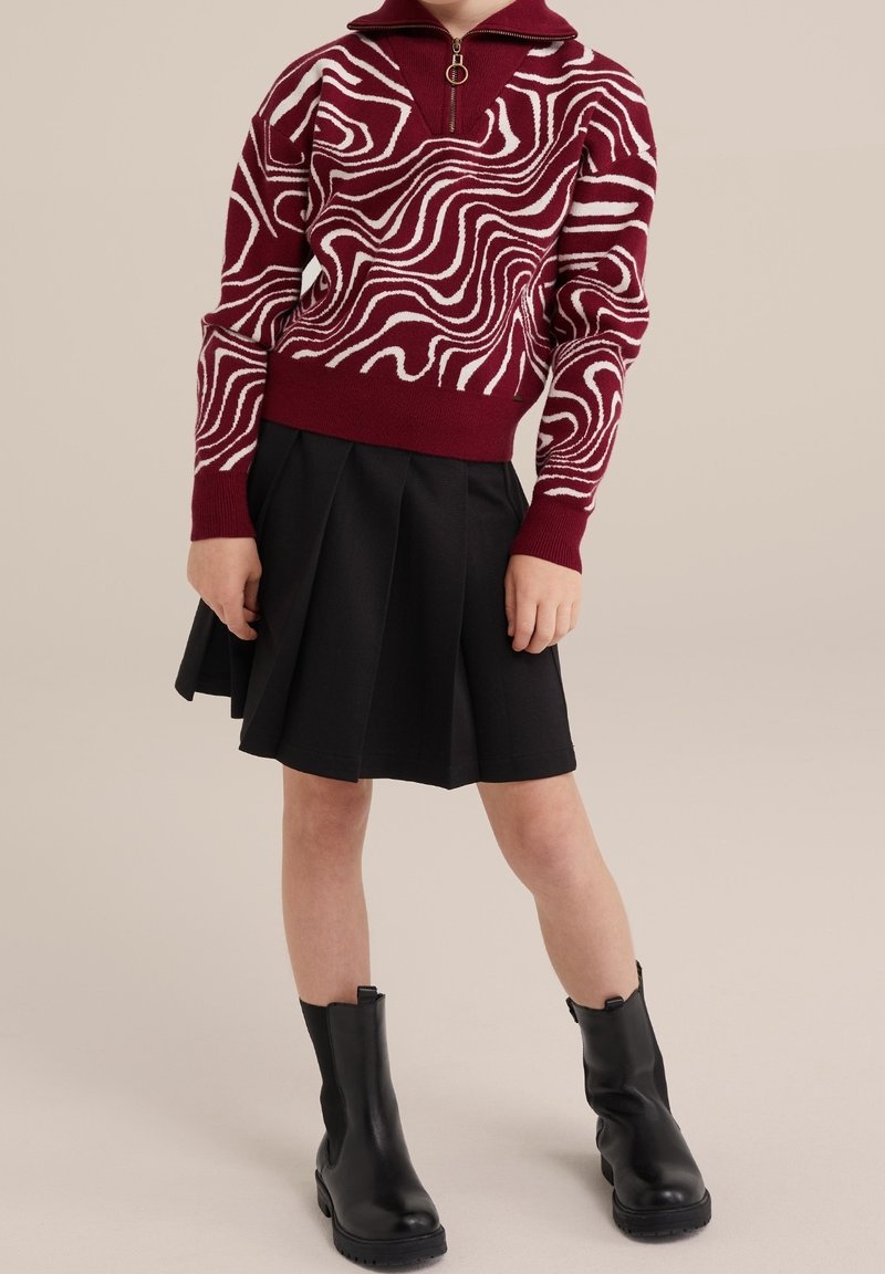 Enfant portant un pull bordeaux et blanc à motifs, une jupe plissée noire et des bottines noires, debout sur un fond beige.
