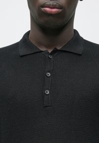 Polo preta em malha com padrão texturizado, contendo colarinho e pala com três botões. O tecido aparenta ser macio e respirável.