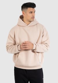Sweatshirt à capuche beige clair avec une poche kangourou, des poignets côtelés et un détail graphique blanc subtil sur la manche gauche. Texture douce.