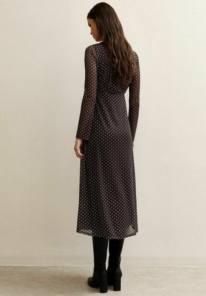 Femme vue de dos portant une robe midi noire à manches longues avec des pois blancs et des bottes à talons noires, devant un mur uni.