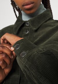Manšestrová bunda tmavě zelené barvy s kovovými patentkami, žebrovanou texturou a límcem. Záběr zblízka na rukáv a detaily látky.