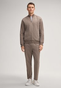 Braune, strukturierte Zip-Jacke mit passender Hose, die dezente Muster aufweist, sowie gerippte Bündchen und weiße Sneakers für einen abgestimmten Look.