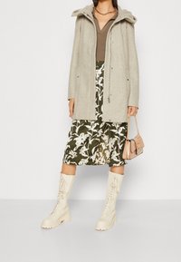 Cappotto grigio chiaro con cappuccio, top marrone, gonna oliva con motivo floreale bianco, stivali al ginocchio color crema con lacci, e una piccola borsa a tracolla beige.